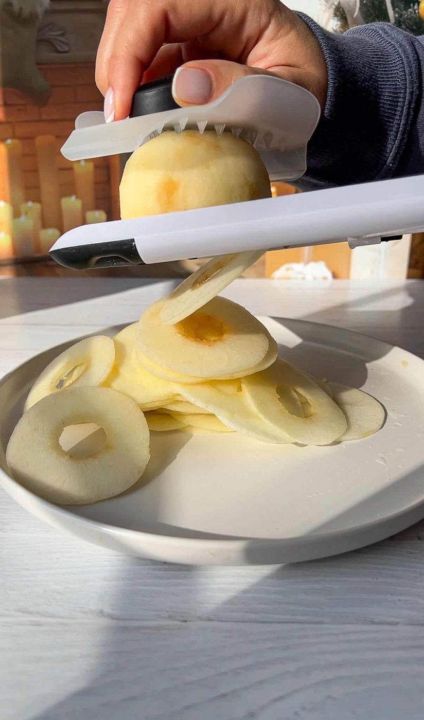 A hand uses a slicer to cut the fruit into thin, uniform rings on a white plate. Getting the apples prepared for the Invisible Apple Cake recipe. 