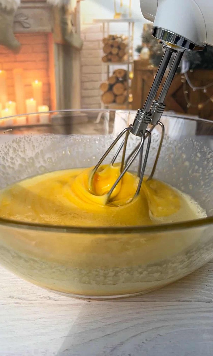 Close-up of an electric mixer beating eggs in a glass bowl. In the background, a cozy room with lit candles, stacked firewood, and a fireplace creates a warm ambiance.