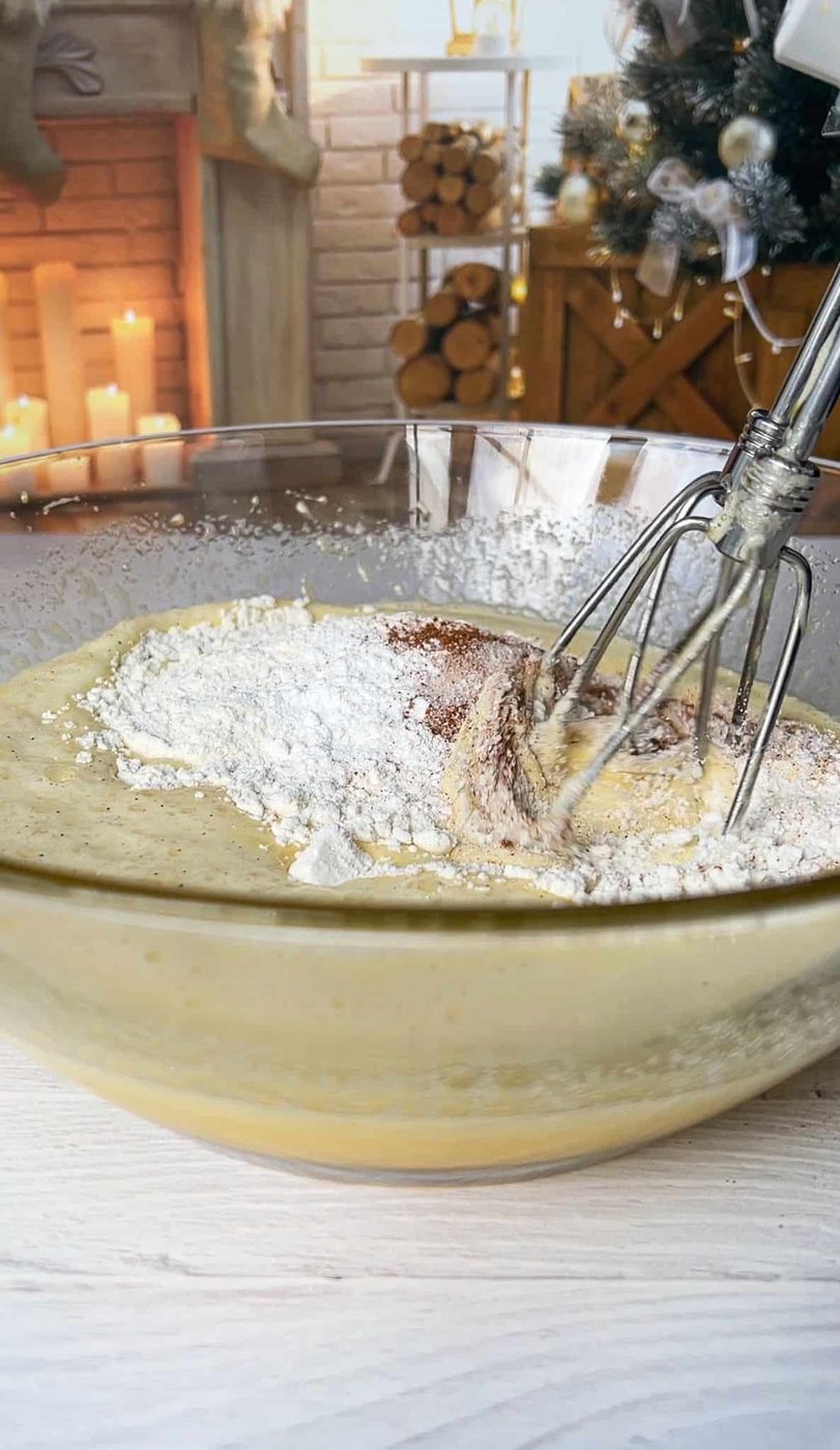 A glass bowl with cake batter being mixed using a hand mixer, next to a fireplace decorated with stockings and candles. A Christmas tree with ornaments is visible in the background.