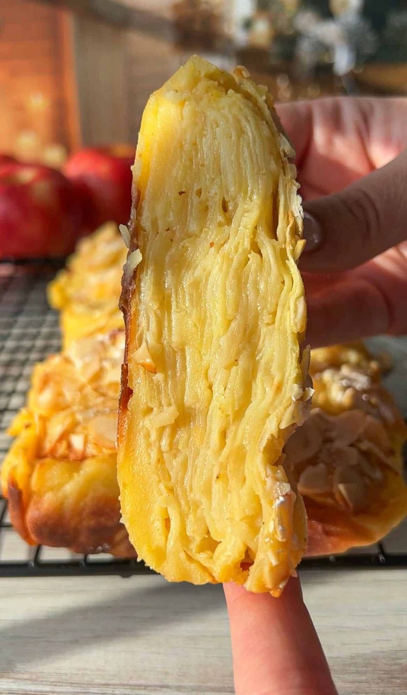 Close-up of a hand holding a slice of Invisible Apple Cake, showcasing the intricate layers within. The dessert has a golden crust, and sliced almonds are sprinkled on top. A cooling rack and red apples are visible in the blurred background.