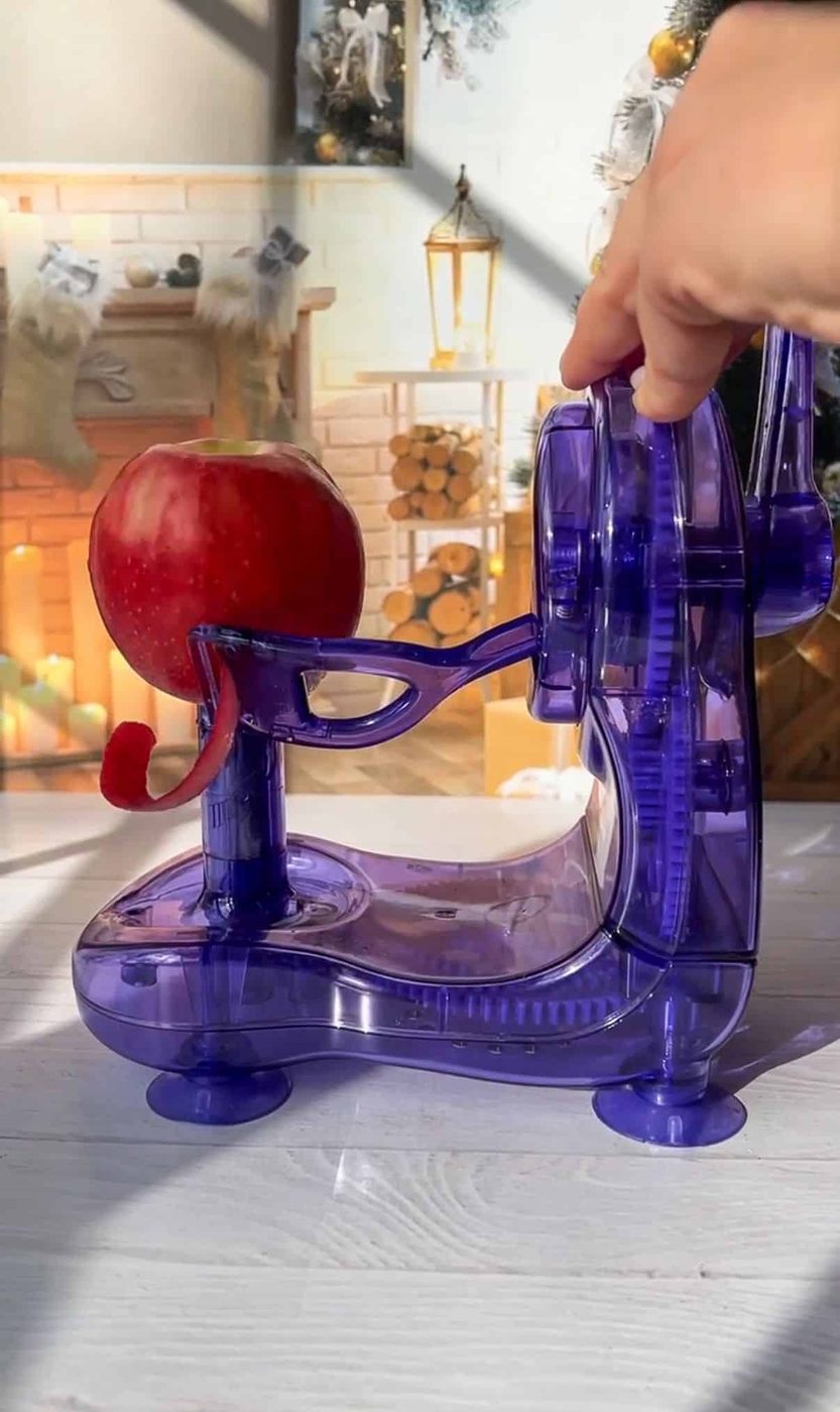 A hand operates a purple apple peeler, peeling an apple. In the background, there's a cozy room setting with a fireplace and a stack of firewood. Sunlight brightens the scene.