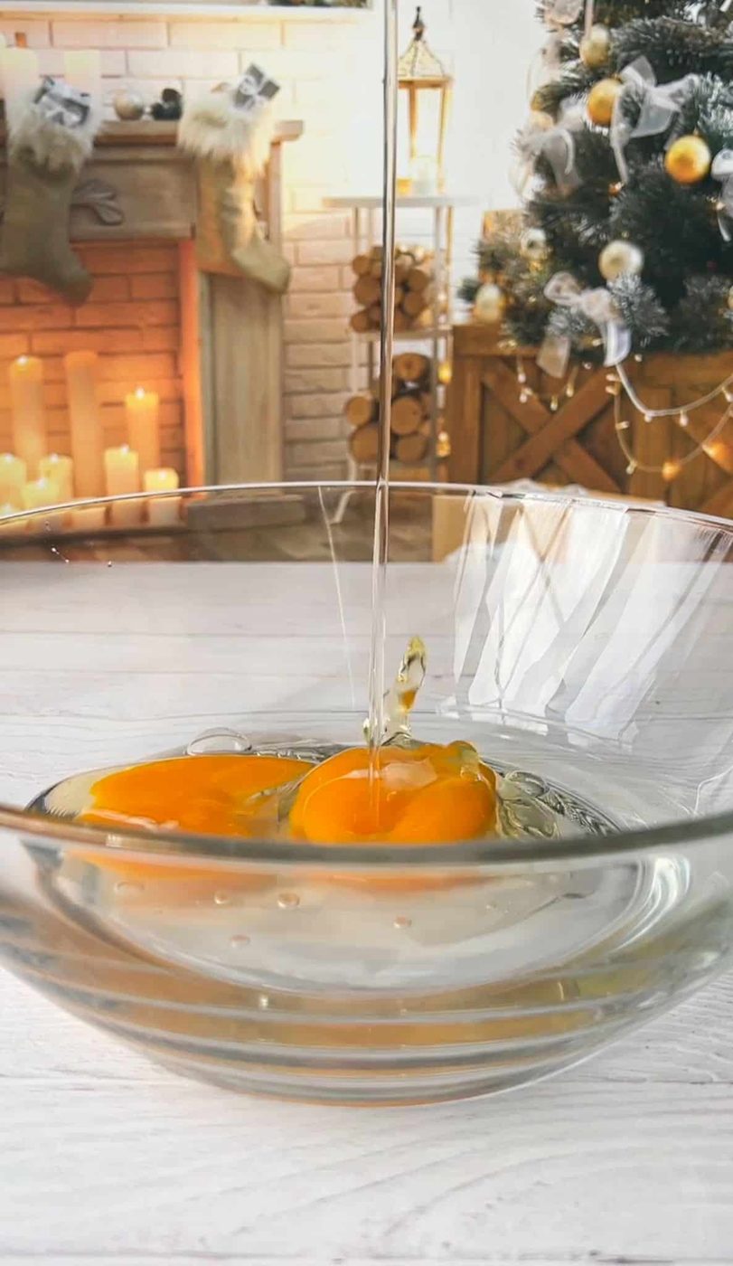 A bowl with cracked eggs is on a kitchen counter. In the background, there's a decorated Christmas tree, a cozy fireplace with stockings, and stacked logs. Candles add a warm glow to the festive scene.
