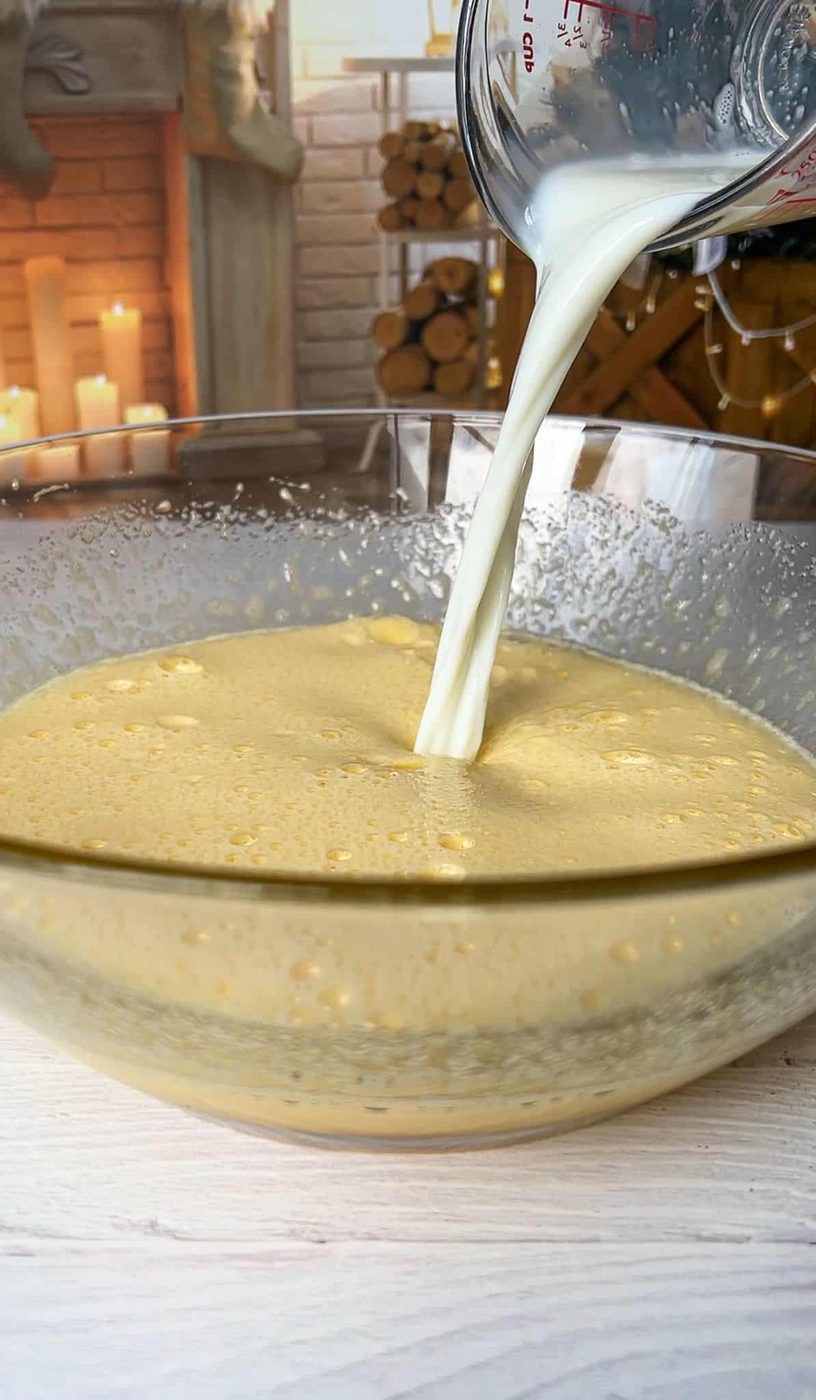 Milk being poured into a glass bowl filled with a frothy yellow mixture. In the background, a rustic setting features stacked firewood and lit candles beside a brick fireplace.
