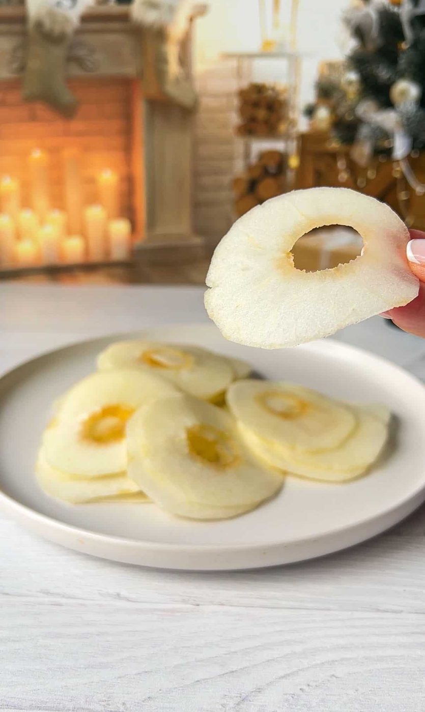 A hand holding the thinly sliced fruit, fireplace and a decorated Christmas tree is in the background. The apples are sliced so thinly that they will disappear into the Invisible Apple Cake. 