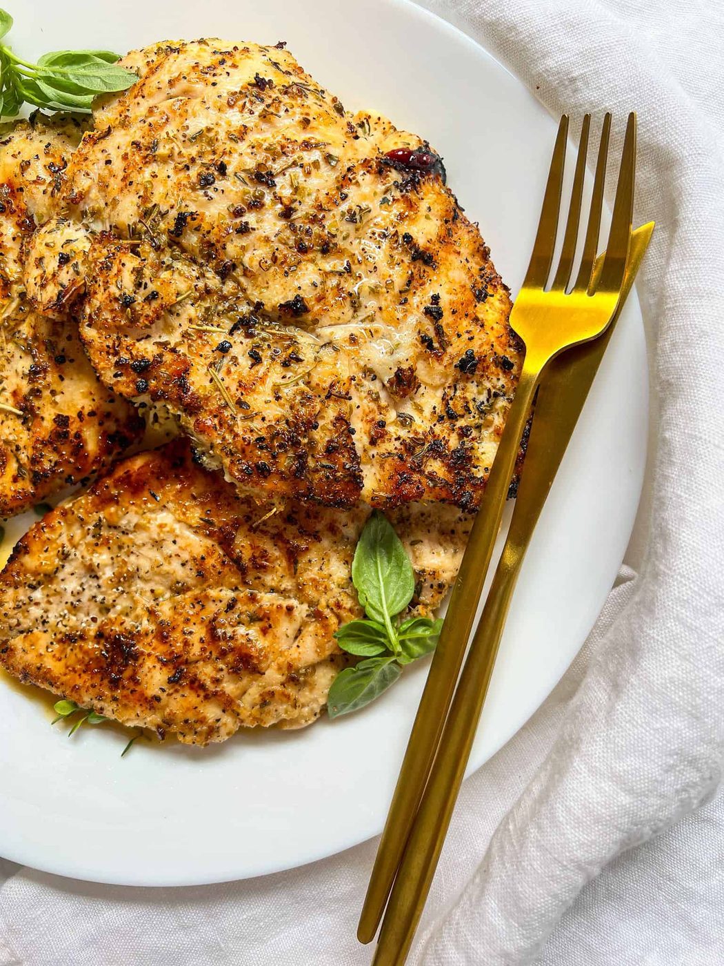Grilled, seasoned chicken breasts on a white plate garnished with fresh basil, next to a gold fork and knife, set on a white cloth background.