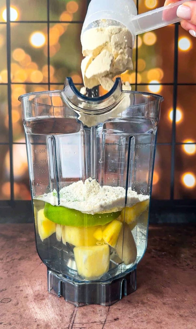A blender is filled with water, pieces of pineapple, and banana. A hand is pouring protein powder from a scoop into the blender. The background is blurred with warm lights, creating a cozy atmosphere.