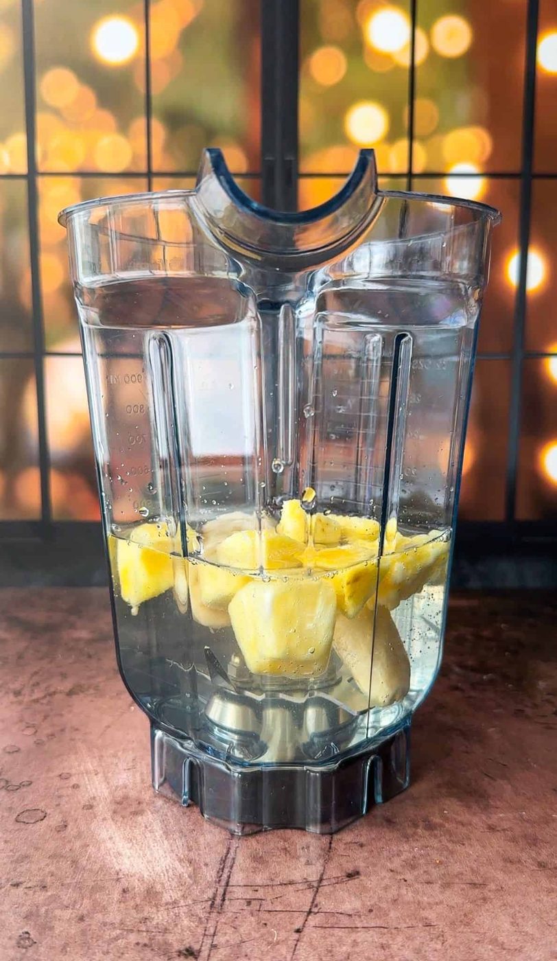 A blender container filled with chunks of pineapple and water sits on a wooden surface. The background features a grid window with warm, circular lights blurred, creating a cozy atmosphere.