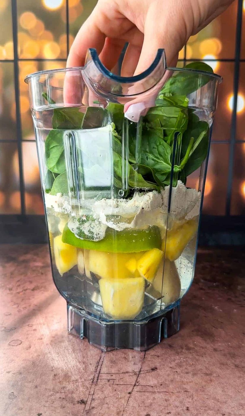 Hand placing spinach leaves into a blender filled with pineapple chunks, lime slices, and protein powder, all ready for blending. The background features soft, warm lighting with a wood-toned surface.