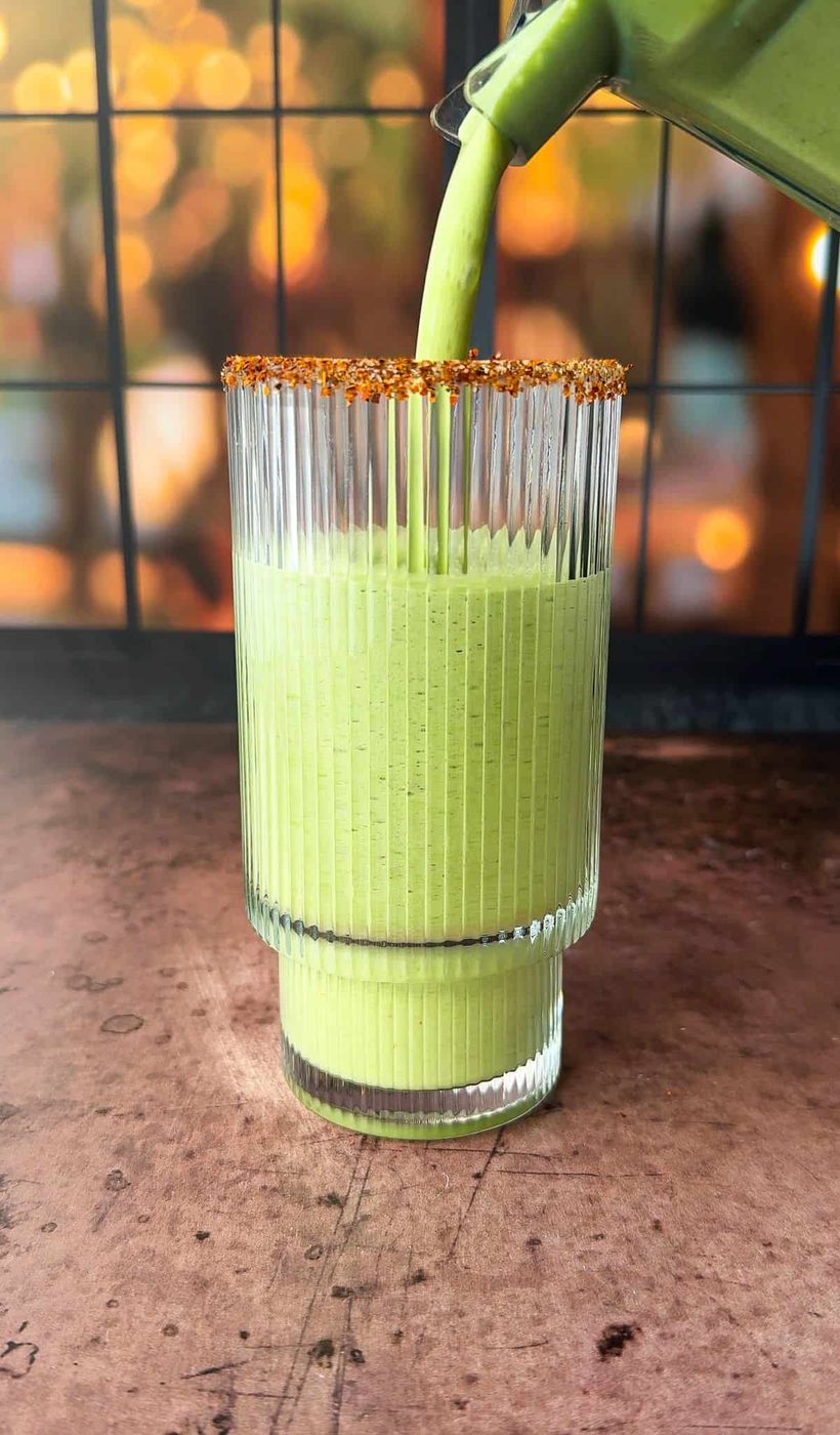 A tall, ridged glass is being filled with a key lime pie smoothie. The rim of the glass is coated with a spice mix. The background is softly blurred with warm, golden hues, suggesting evening light through a window.