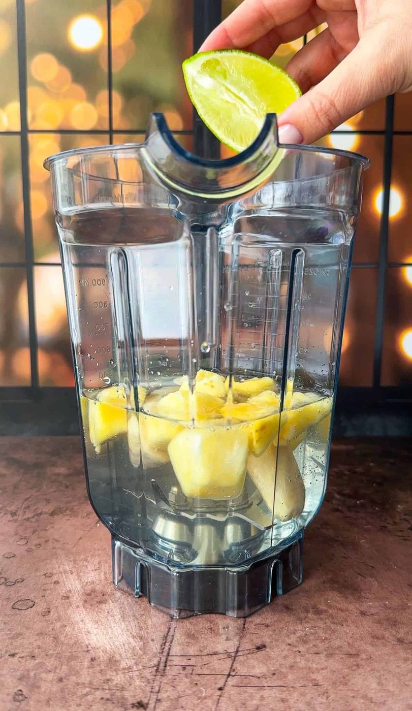 A blender container filled with pineapple chunks, banana and water. A hand is squeezing a lime wedge above the container. The background features soft, bokeh-style lights and a grid-patterned window.