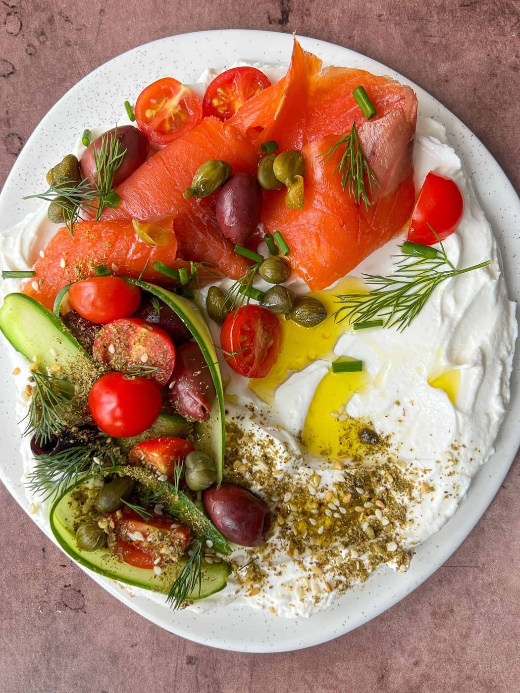 A white plate with smoked salmon, creamy labneh, cherry tomatoes, cucumber slices, olives, capers, and fresh dill. The dish is garnished with olive oil and za'atar seasoning, set on a rustic brown surface.