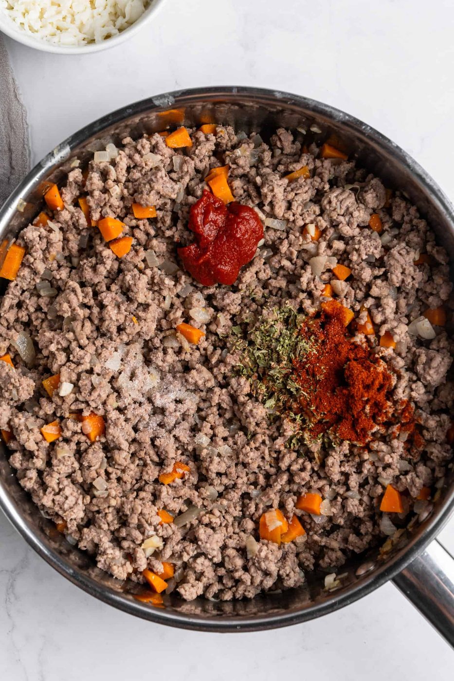 A pan containing browned ground beef mixed with chopped onions and carrots. There's a dollop of tomato paste and sprinkled spices, including paprika and dried herbs, on top. The scene is set on a marble countertop.