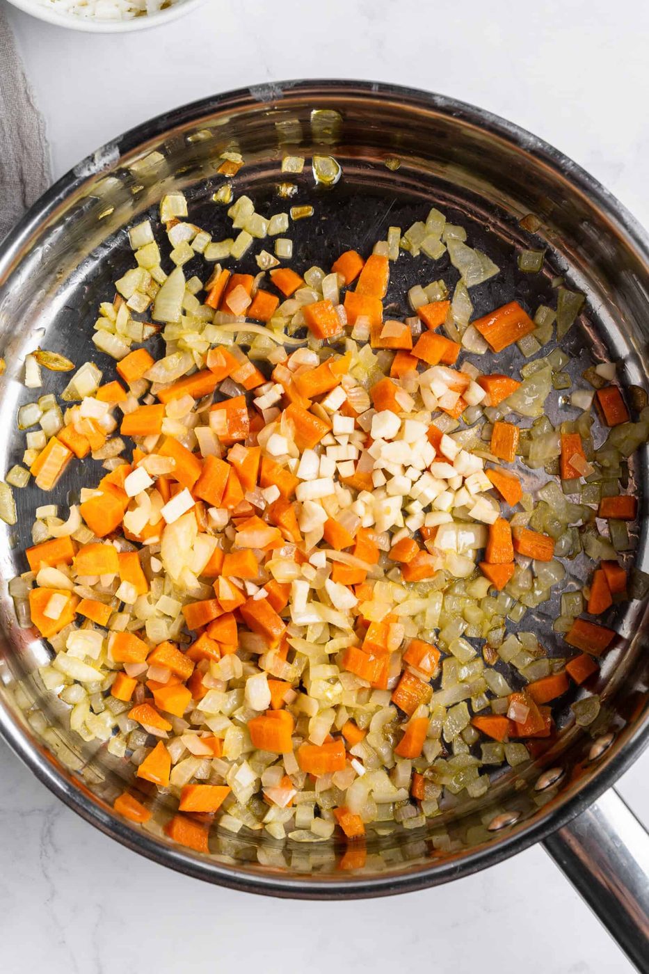 A skillet containing diced onions, carrots, and garlic sautéed until softened and lightly golden. The vegetables are evenly distributed across the shiny surface of the pan, with a few flecks of green herbs visible. The background is a white countertop.