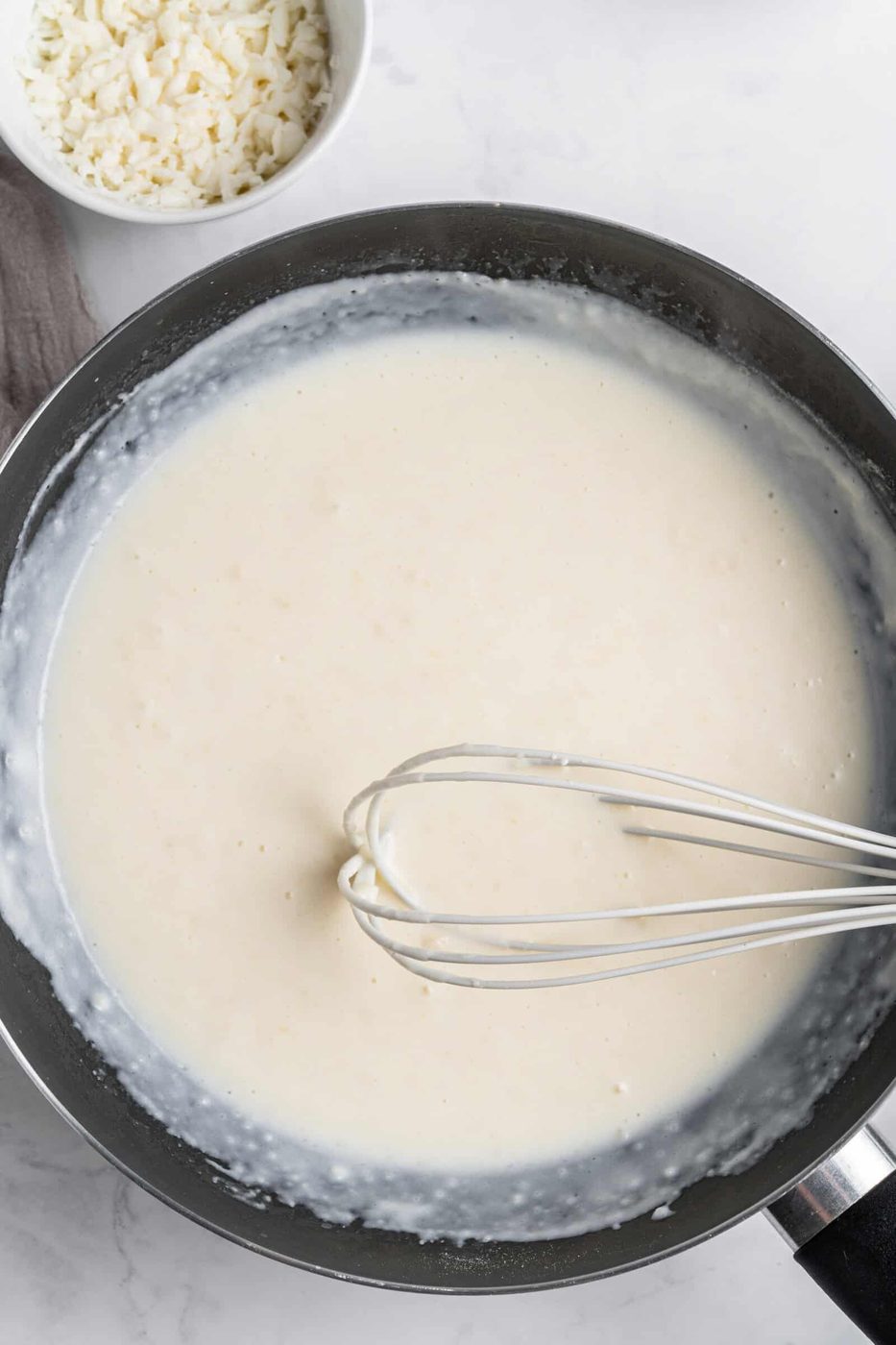 A whisk stirring a creamy white sauce in a black pan on a marble surface. A small white bowl containing grated cheese is placed nearby.