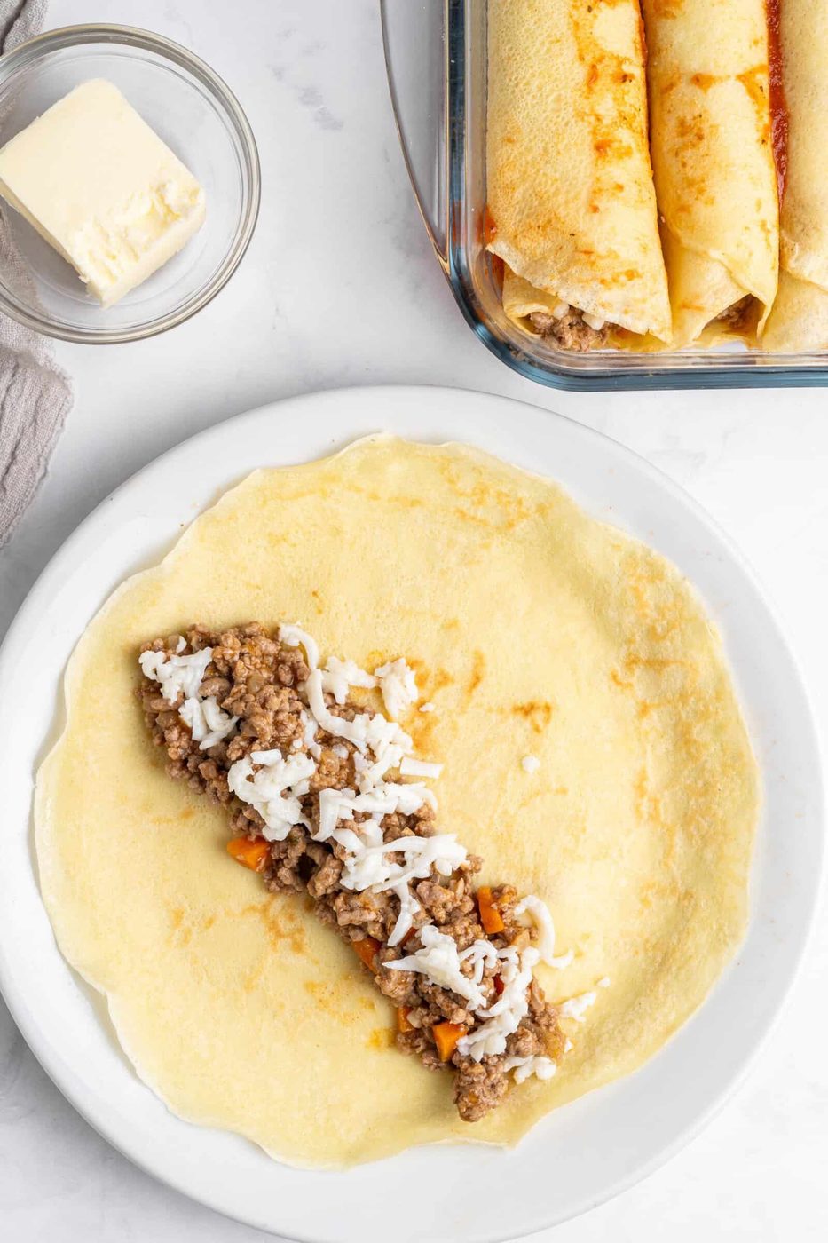 A flat pancake on a white plate is filled with seasoned ground meat and shredded cheese, ready to be rolled. Nearby, a glass dish holds two rolled pancakes. A small bowl of butter sits beside a folded napkin on a marble surface.