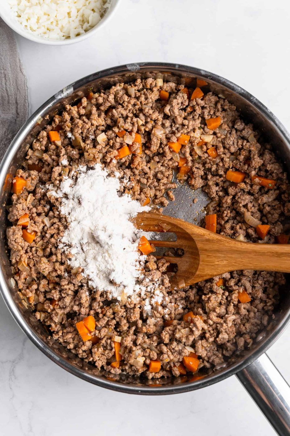 A skillet filled with cooked ground meat and chopped orange bell peppers, topped with a pile of flour. A wooden spatula rests inside the skillet, and a small bowl of white shredded cheese is visible in the background.