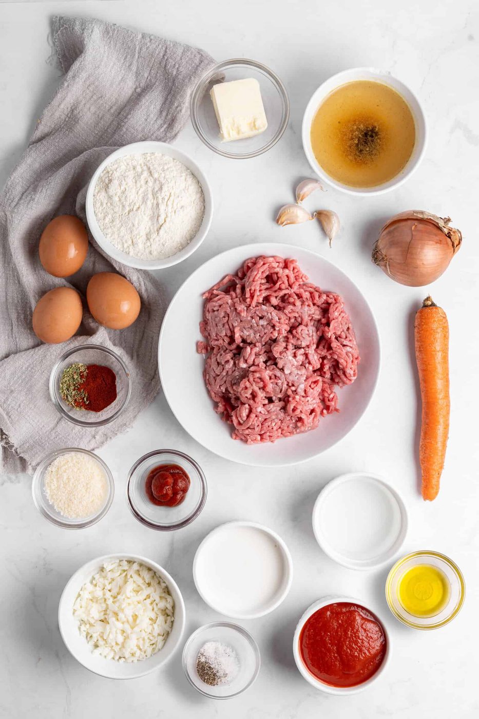 Top-down view of various ingredients on a marble surface, including ground meat, eggs, flour, butter, stock, garlic, onion, carrot, spices, tomato sauce, and oil, all arranged in bowls and on a linen cloth.