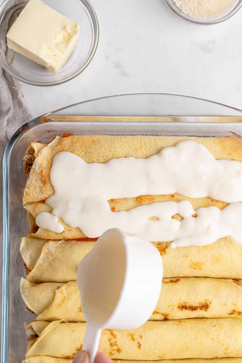 A baking dish filled with rolled crepes is being topped with a creamy sauce. A hand is pouring sauce from a measuring cup. Nearby, there is a dish of butter and a bowl of grated cheese.