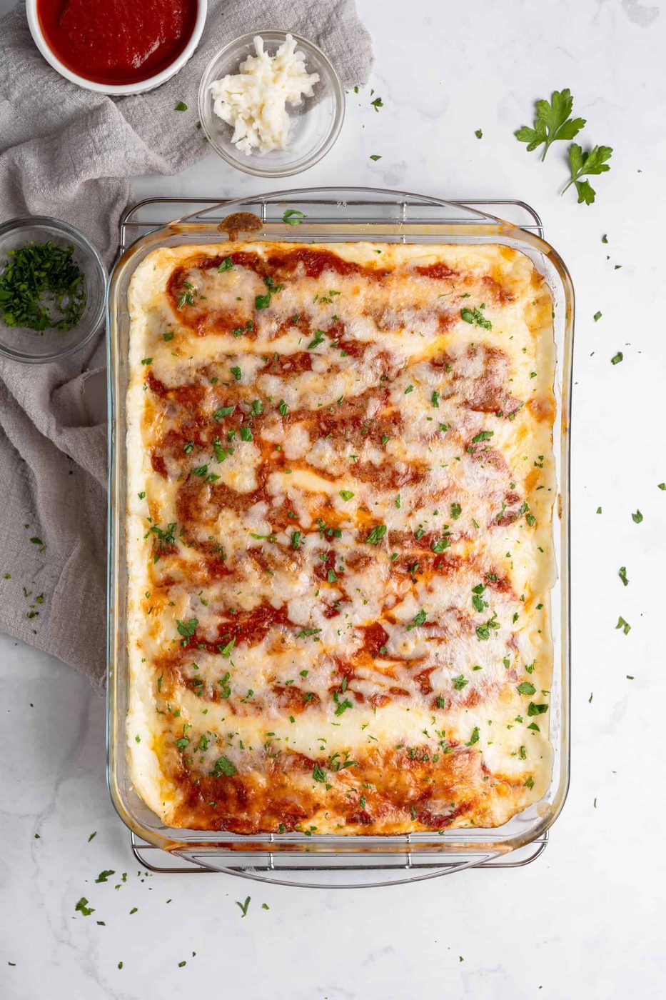A glass baking dish filled with freshly baked lasagna topped with melted cheese and sprinkled with chopped parsley. Ingredients like grated cheese and tomato sauce are nearby on a white marble surface.