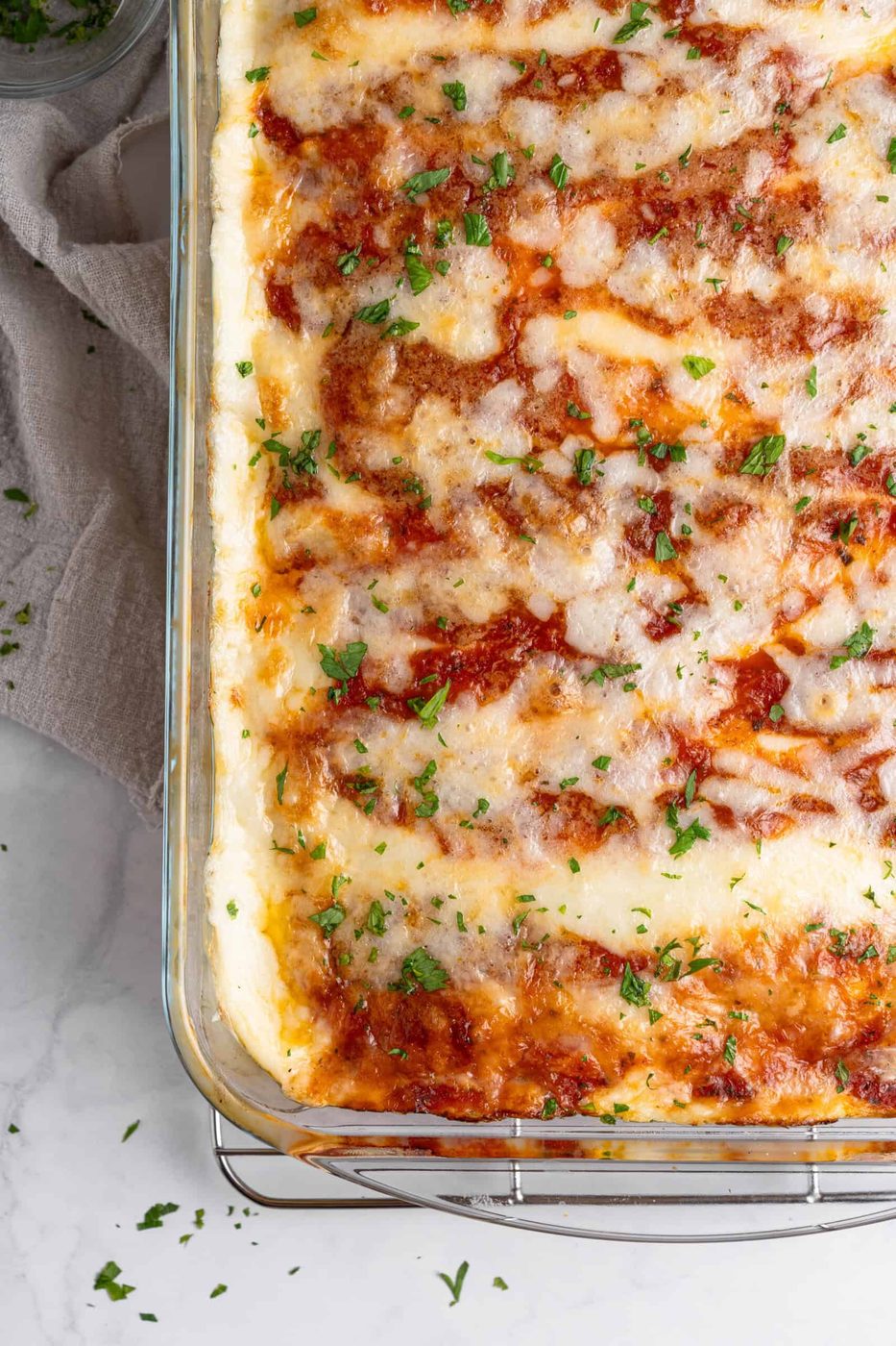 A close-up of a freshly baked tray of lasagna with crepes in a glass dish, topped with melted cheese and sprinkled with chopped parsley. The lasagna is resting on a cooling rack, and a linen cloth is visible on the side.