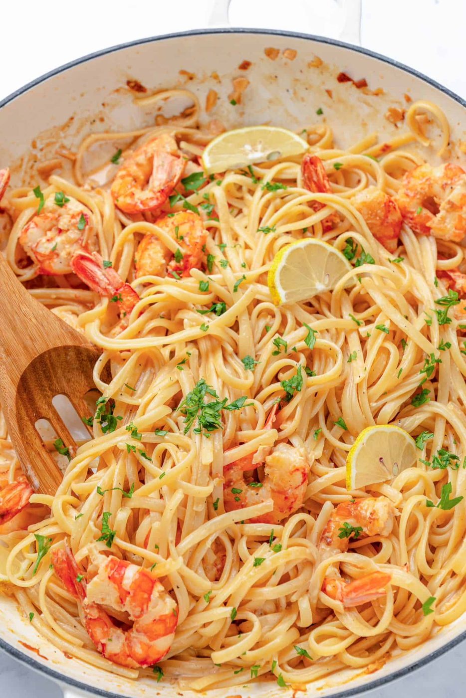 A dish of shrimp linguine is garnished with lemon slices and parsley. The pasta is mixed with succulent shrimp, and a wooden spoon is visible in the creamy sauce.
