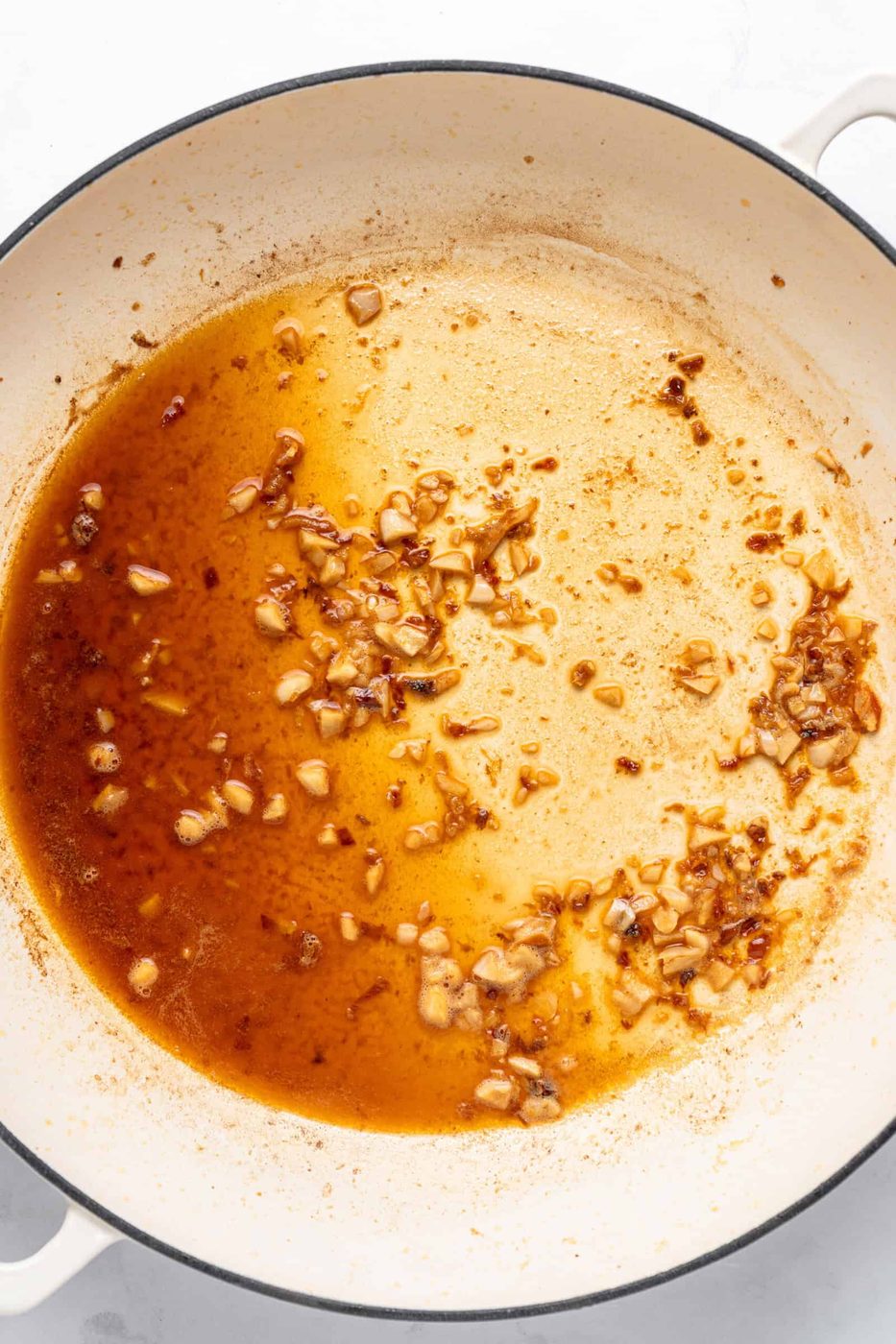 A white pan with browned garlic pieces in a small amount of oil, creating a golden-brown mixture. The garlic appears slightly caramelized and the pan shows some darker spots from cooking.
