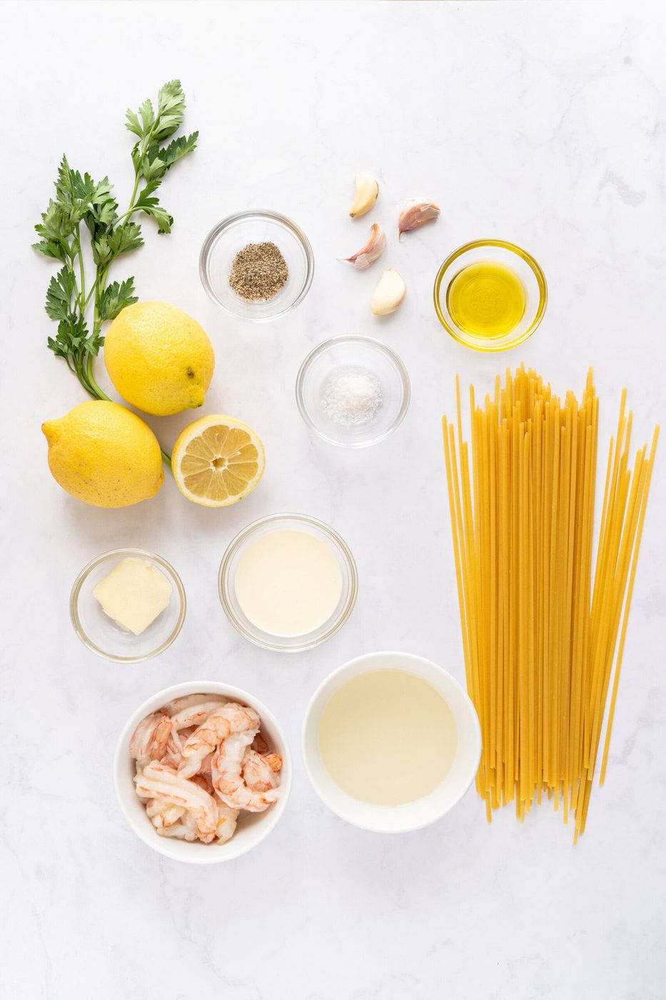 Ingredients for a pasta dish are arranged on a white surface, including parsley, lemons, garlic, olive oil, pepper, salt, butter, cream, shrimp, white wine, and uncooked pasta.