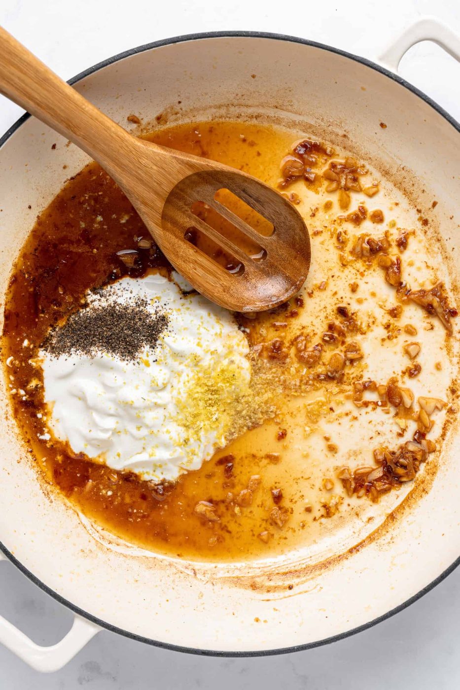 A white pan containing a wooden spoon, sautéed onions, seasonings, and a dollop of yogurt being mixed. The spices include black pepper and lemon zest, creating a rich, aromatic sauce base.