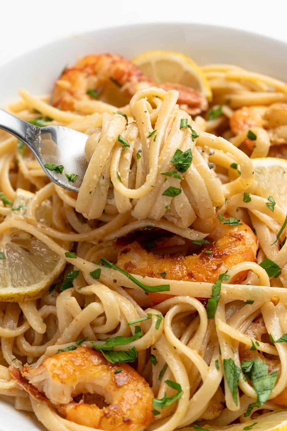 A close-up of a fork twirling linguine pasta with shrimp. The pasta is garnished with parsley and lemon slices, showcasing a creamy sauce. The shrimp are golden and cooked, adding texture and color to the dish.