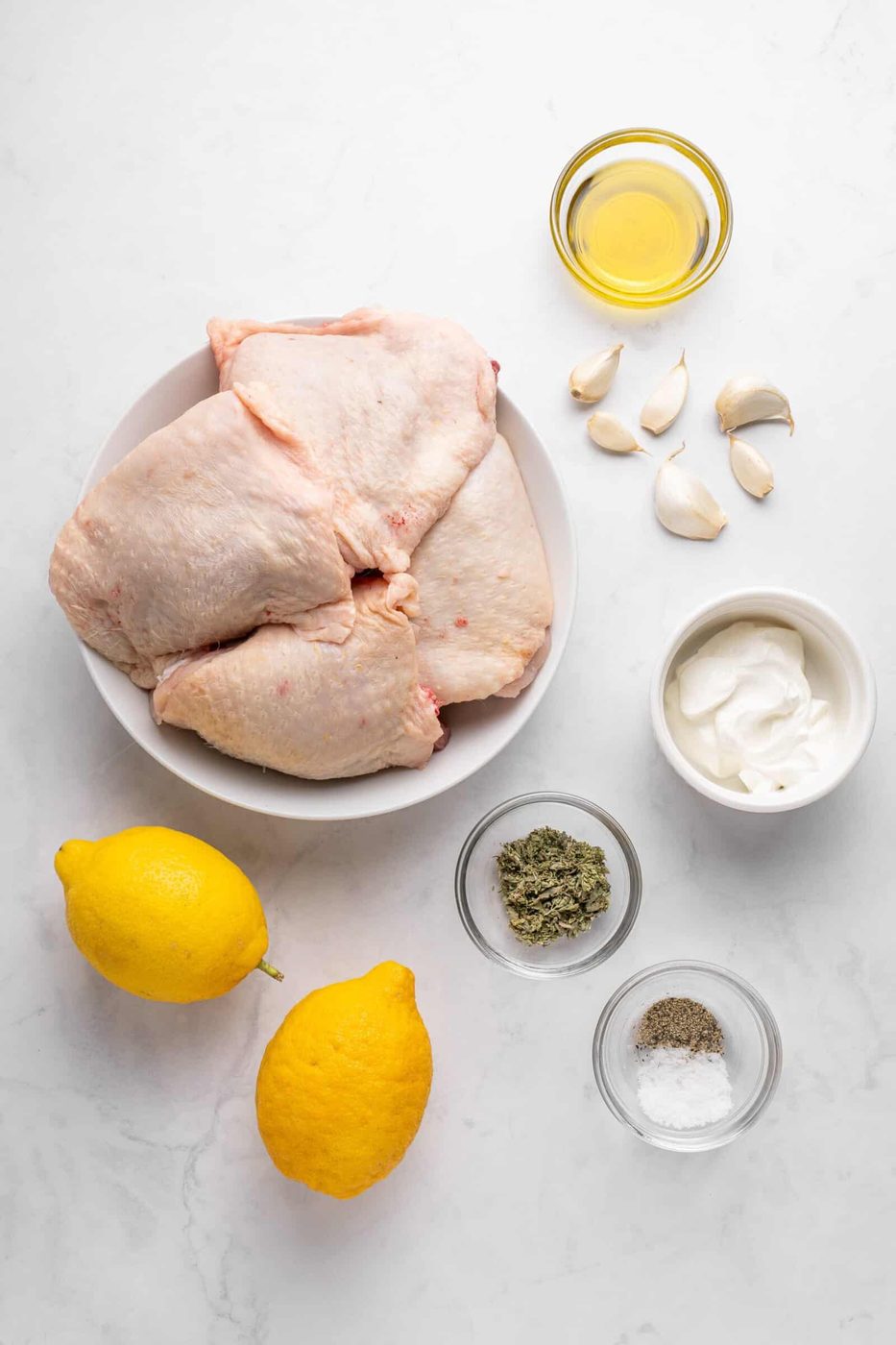 A flat lay of raw chicken thighs in a bowl, surrounded by garlic cloves, a small bowl of olive oil, two lemons, a bowl of yogurt, herb seasoning, salt, and pepper, all on a white marble surface.