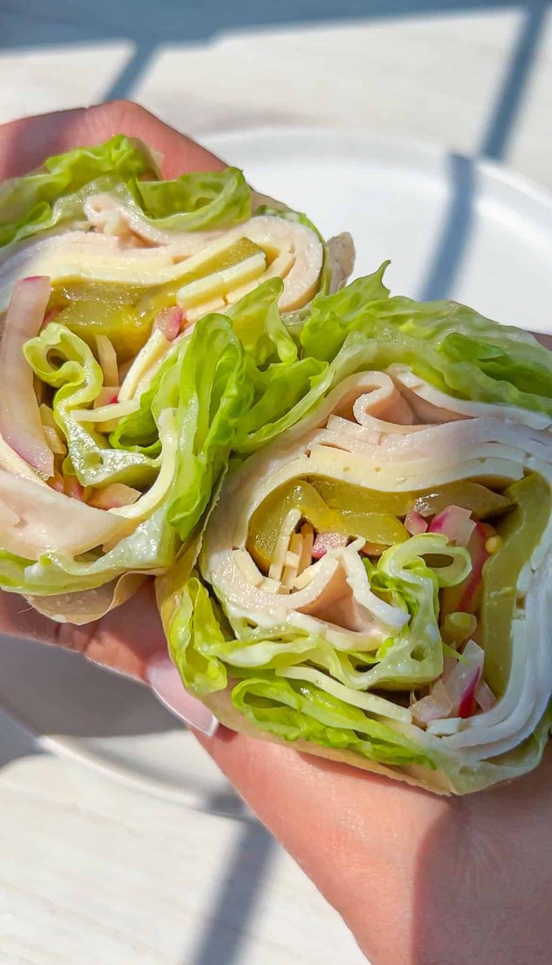 Two hands hold a sandwich wrapped in fresh lettuce leaves, filled with slices of turkey, cheese, pickles, onions, and other vegetables, instead of bread.