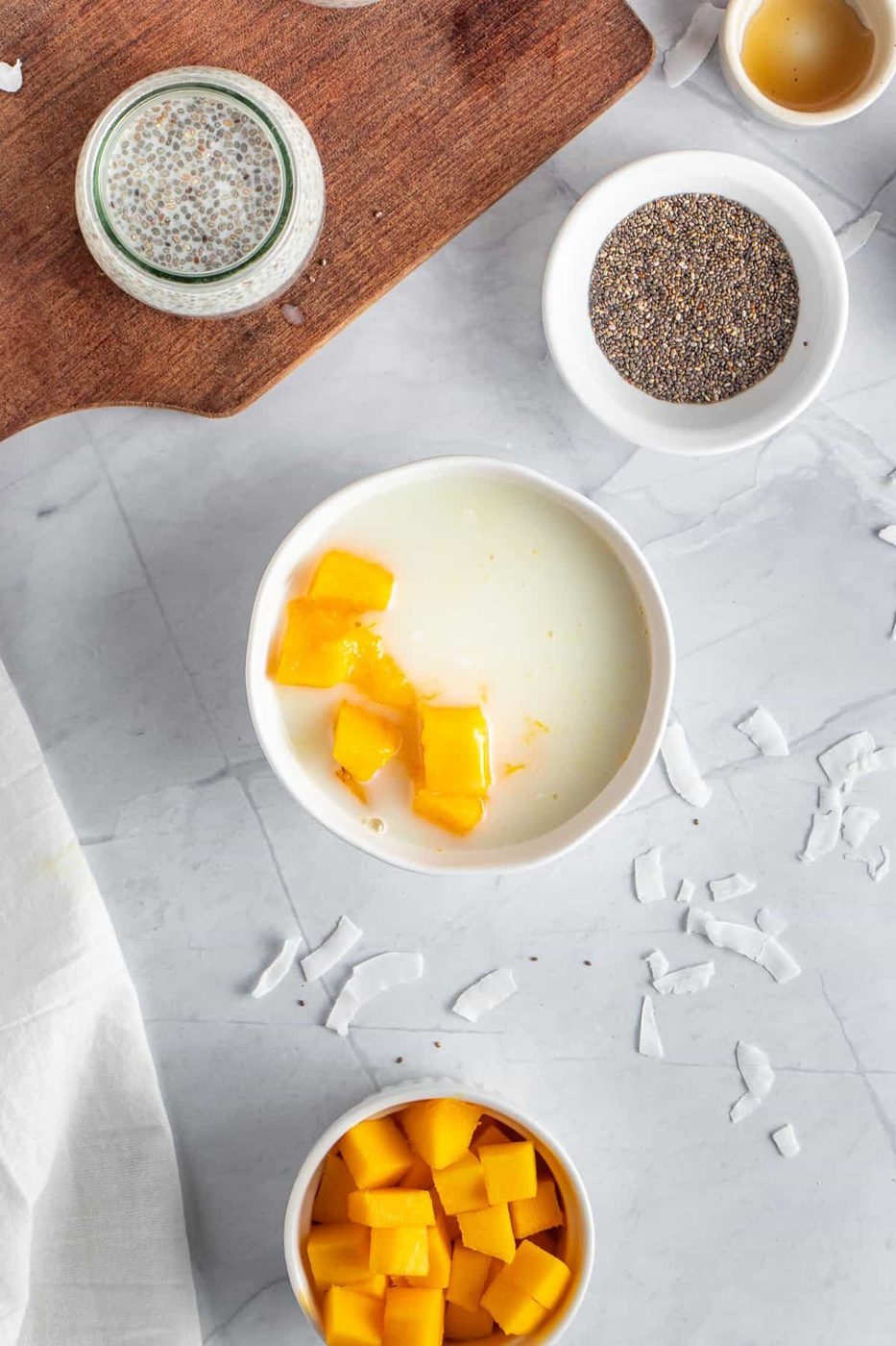 A white bowl of yogurt topped with mango chunks on a marble surface. Nearby are small bowls containing chia seeds, and diced mango. Coconut flakes and a wooden board are also visible.