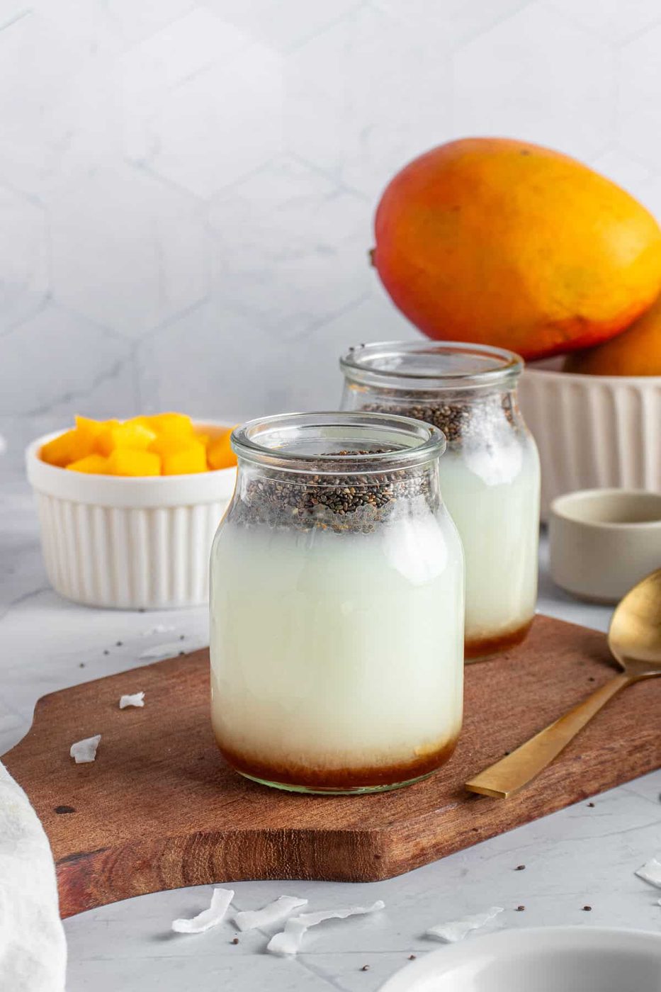 Two glass jars of a chia seed pudding mixture sit on a wooden board. Fresh mango chunks are in a small white bowl, with a whole mango nearby. A gold spoon rests on the board, and a light, hexagonal-tiled background completes the scene.