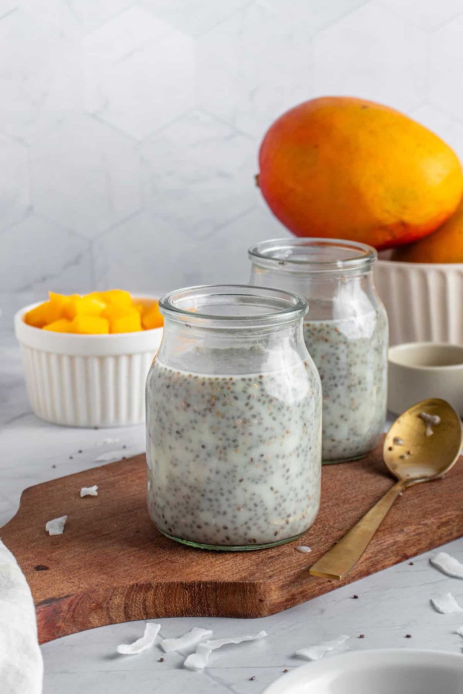 Two glass jars filled with the pudding mixture on a wooden board. A golden spoon rests beside them. In the background, a bowl of diced mango and two whole mangoes are visible. The setting is a marble surface with a subtle hexagonal pattern wall.