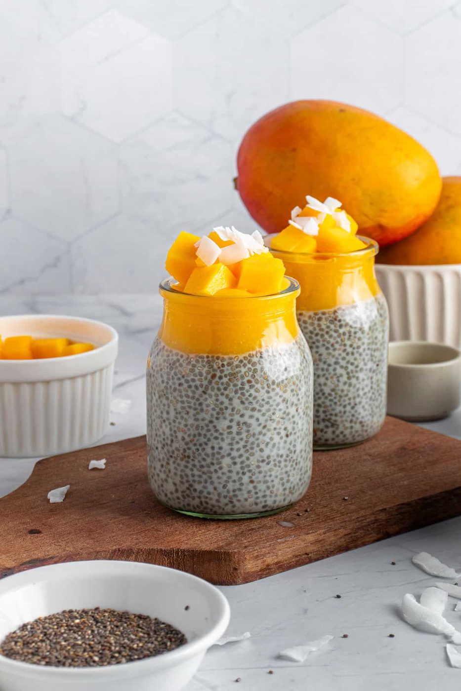 Two jars topped with diced mango and coconut flakes sit on a wooden board. A bowl of chia seeds and mango pieces are nearby. Whole mangoes are in the background against a light-colored geometric tile wall.