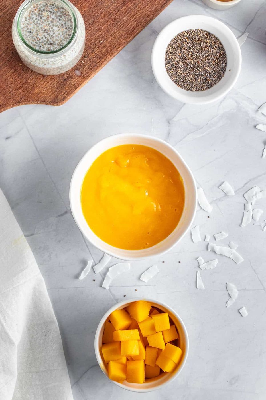 Overhead view of a smoothie bowl with bright yellow mango puree in a white bowl, surrounded by bowls of chia seeds, diced mango, and coconut flakes on a marble countertop. A cutting board with ingredients is partly visible.
