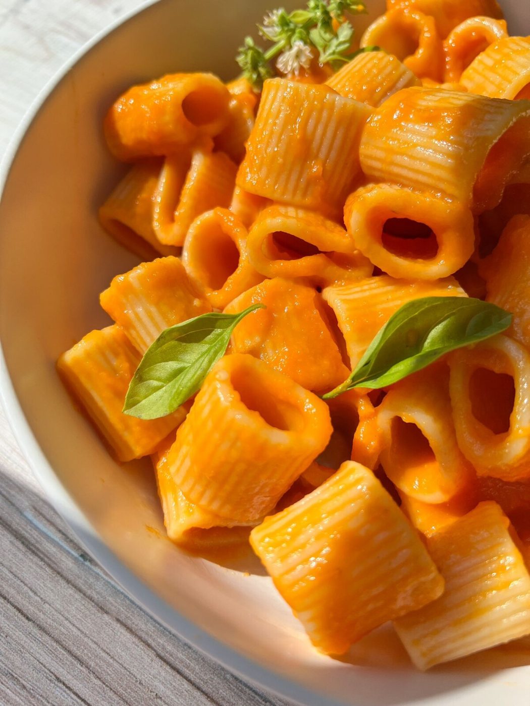 A bowl of rigatoni pasta covered in a vibrant orange Marinara Sauce, garnished with fresh basil leaves and a sprig of thyme. The pasta is well-coated and appears appetizing, with the sunlight casting a warm, natural light on the dish.