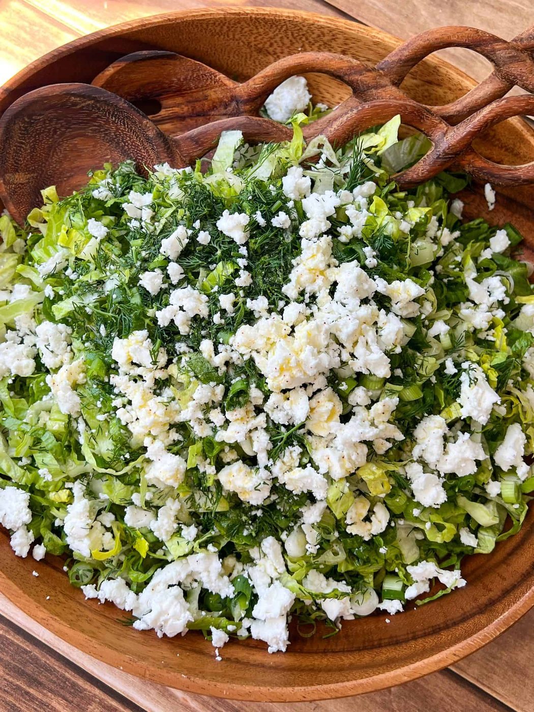 A wooden bowl filled with chopped lettuce, fresh herbs, and crumbled white cheese, with wooden salad servers resting on the edge. The salad sits on a wooden surface.