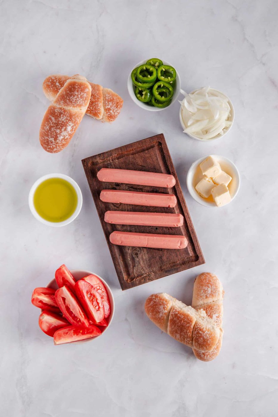 Ingredients for a meal are laid out on a marble surface, featuring hot dog sausages on a wooden tray, sliced green jalapeños, onion slices, cubed cheese, tomato wedges, bread rolls, and a bowl of olive oil.