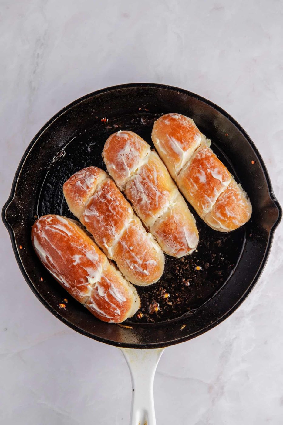 Four golden-brown, oval-shaped buns are in a black skillet, drizzled with a creamy white glaze, on a light marble surface.
