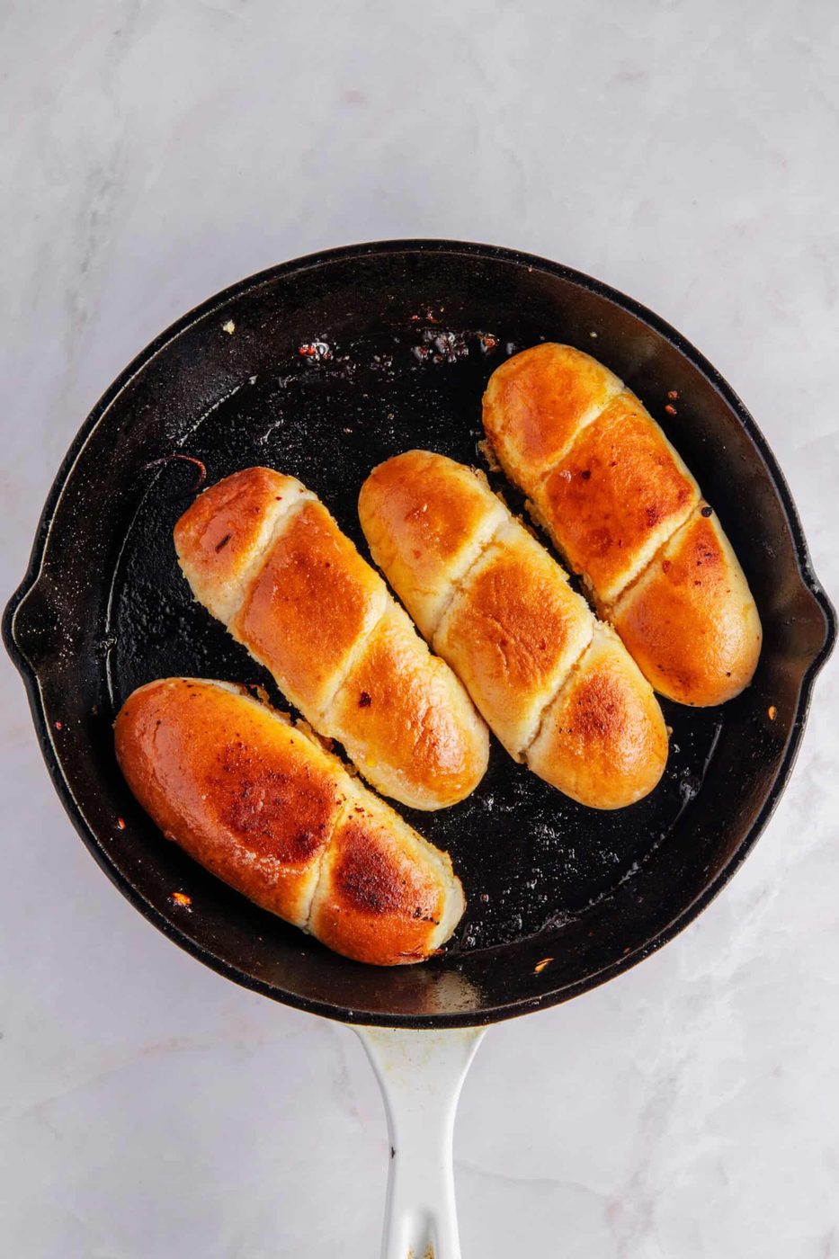 Four golden-brown buns are aligned in a black skillet with a white handle, on a light marble surface. The buns have a shiny, glazed finish and are arranged parallel to each other.