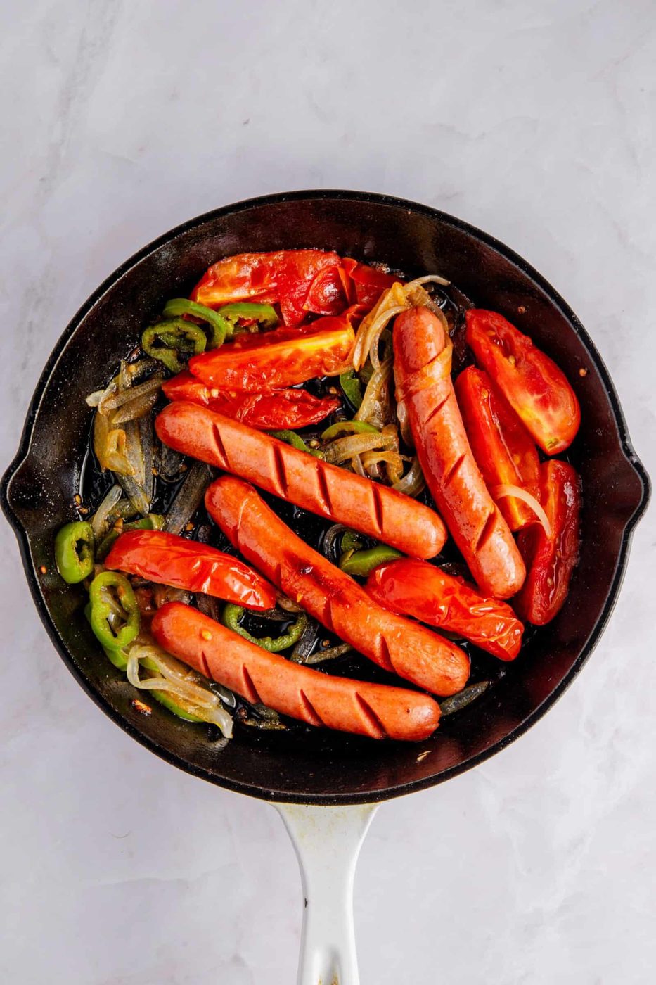 Pan with cooked hot dogs, sliced green peppers, tomatoes, and onions in a skillet on a light marble surface. The hot dogs and vegetables appear grilled and slightly charred.