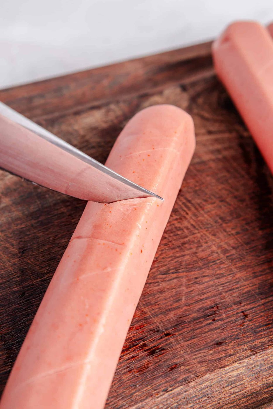 Close-up of a knife slicing into a pink sausage on a dark wooden cutting board. The sausage has light diagonal cuts across its surface. Another sausage is partially visible in the top right corner.