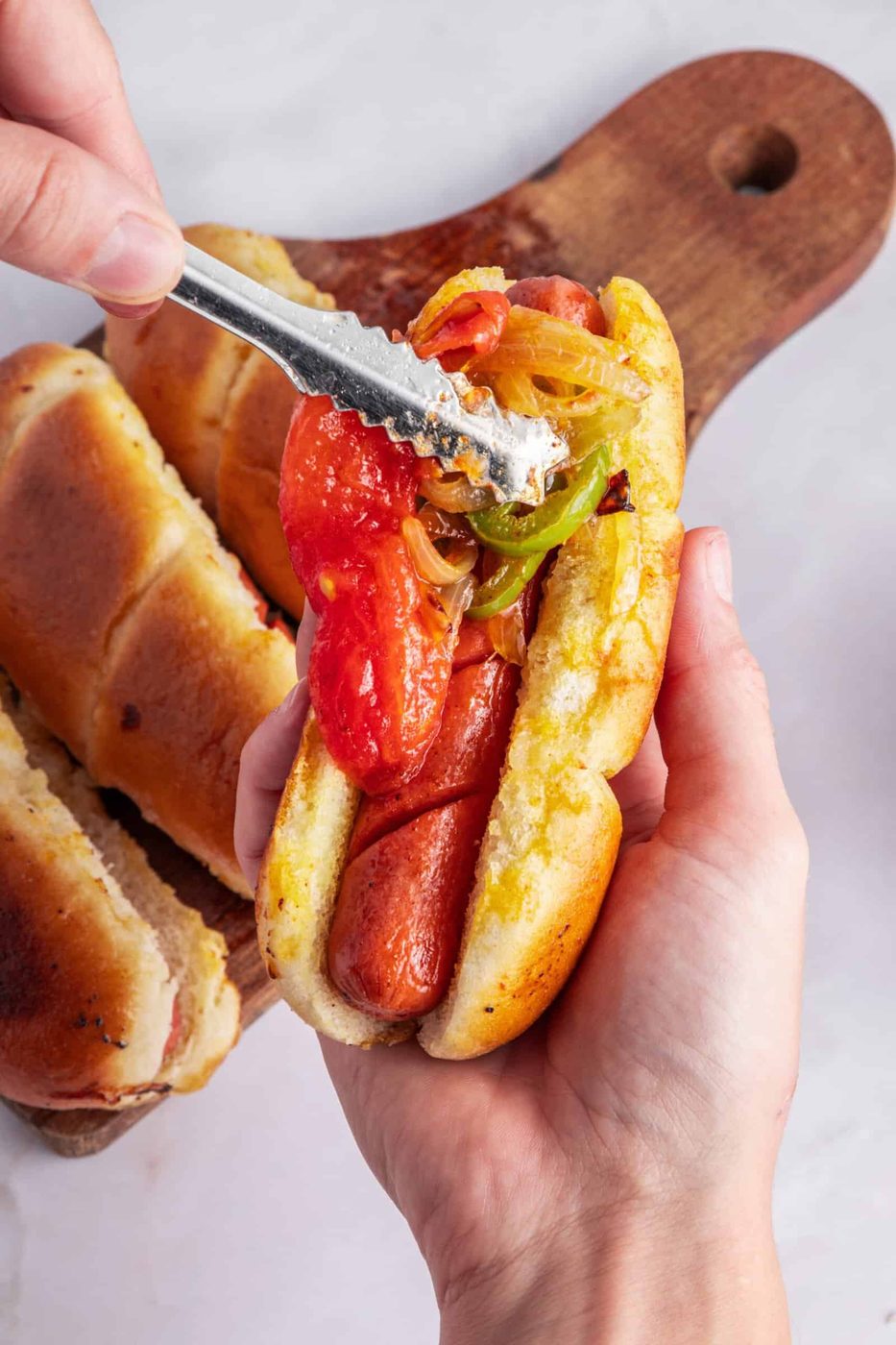 A person holds a hot dog in a bun, topped with sautéed onions and tomato slices. Another hand spreads mayonnaise on the toppings with a knife. Two additional hot dog buns are visible on a wooden board in the background.