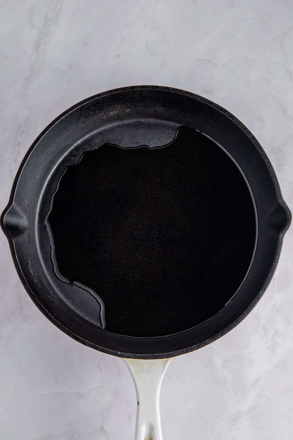 A black frying pan with a white handle on a marble surface, containing a small amount of oil pooling to one side.