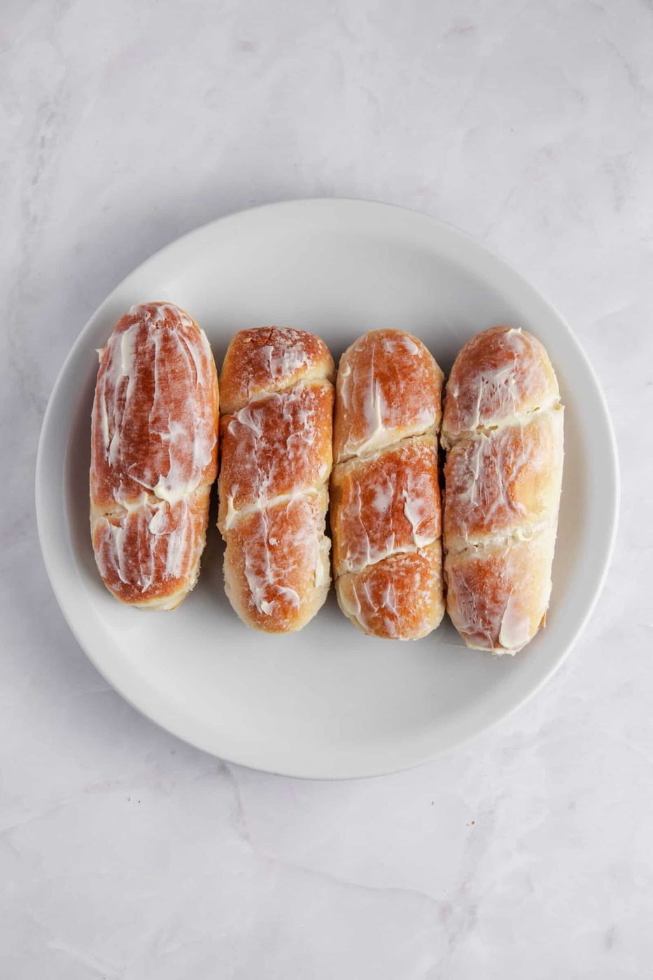 Four glazed pastries are neatly arranged on a white plate. The pastries are covered with a light layer of icing, and they are placed on a white marble surface.