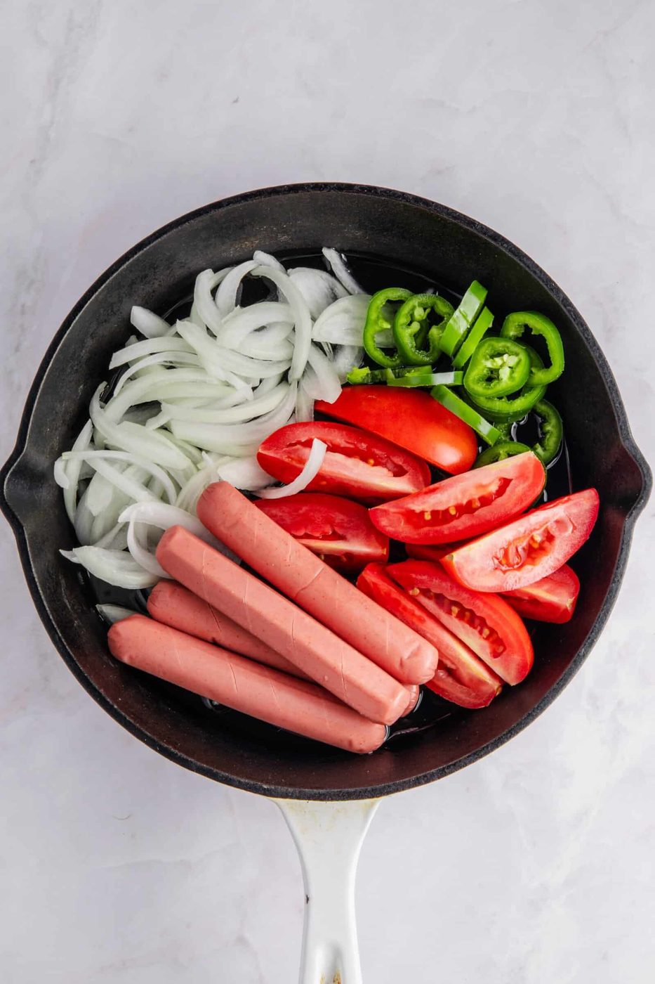 A black pan containing sliced onions, halved tomatoes, sliced green peppers, and four uncooked hot dogs on a white marble surface.