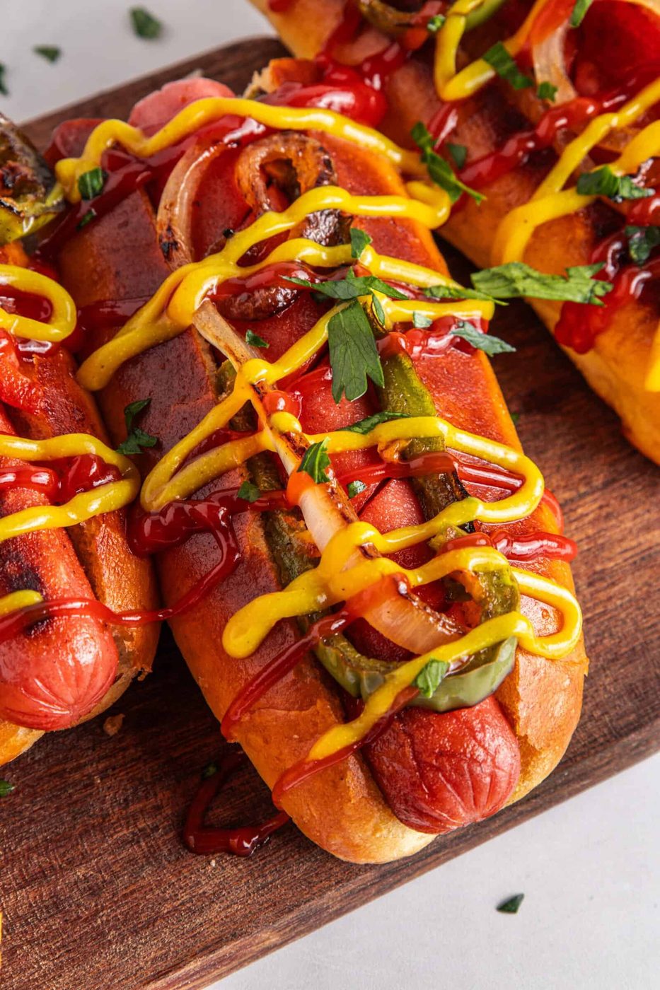 Three grilled Mexican Hot Dogs in buns topped with ketchup, mustard, grilled onions, peppers, and fresh parsley, arranged on a wooden board.