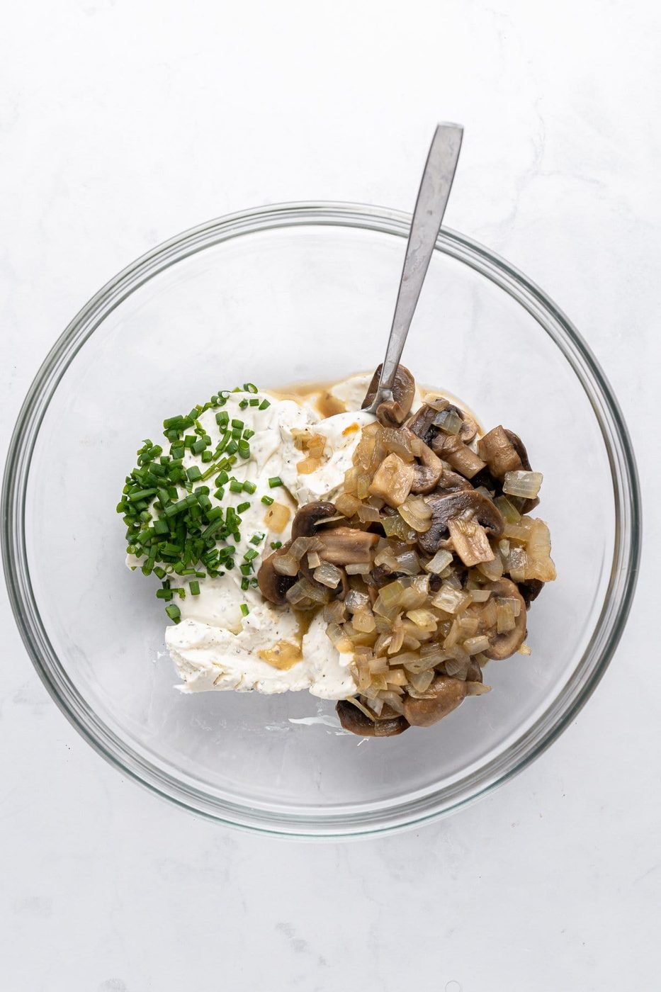 A glass bowl containing a mixture of cream cheese, sautéed mushrooms and onions, and chopped chives. A metal spoon is placed inside the bowl on a light marble surface.