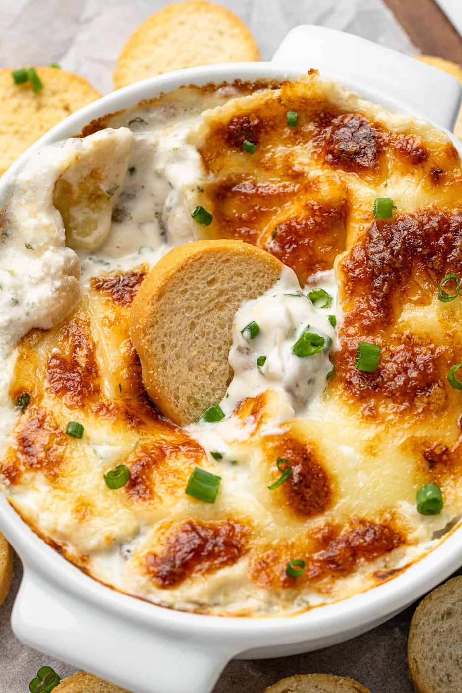 A close-up of a baked cheese dip in a round dish, topped with melted golden-brown cheese and chopped chives. A crispy, round bread slice is dipped in the center. Assorted bread slices surround the dish on a wooden surface.